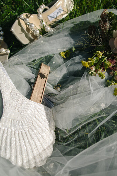 Several bridal items scattered over green grass. A white, beaded high-heeled sandal or platform shoe with pearl accents wrapped around the ankle strap. A wooden hand fan. 