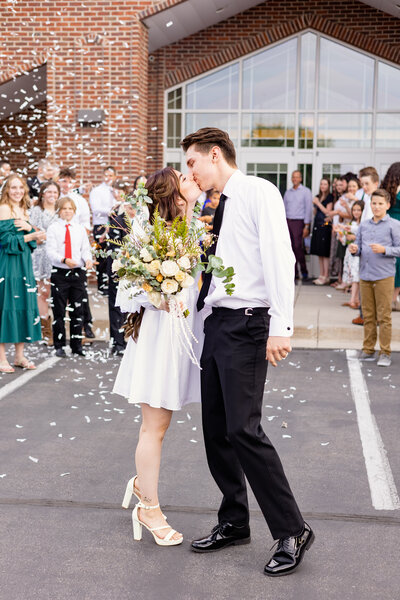 Young bride and groom share one last kiss at their wedding, surrounded by loved ones and confetti.