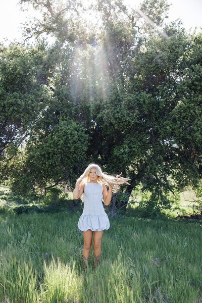 Dreamy backlit senior portrait of a blonde girl twirling in a light blue sundress in a grassy field with sun rays shining through the trees in the background.