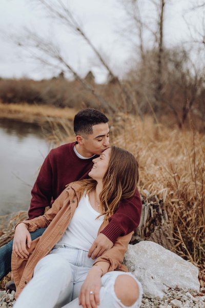 Couple sitting on the ground next to pond. Boy is supporting the girl with his arms around her and kissing her forehead. Both eyes are closed.