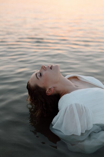 A bride is laying in the water with her hair lightly drenched as the sunset highlights her side profile elegantly.