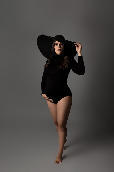 Expectant London, Ontario Mom at a maternity photoshoot in top photography studio. Mom is earing a black collared bodysuit, bare legs, and a wide brimmed felt hat. One had is under her bump, the other hand is gently touching her hat. She is looking at the camera with a half smile.