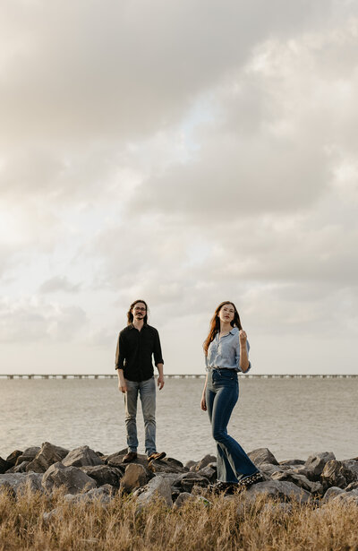 A couple standing on rocky terrain by Lake Pontchartrain, with a bridge in the background under a cloudy sky. The man wears a black shirt and jeans, while the woman is in a light denim shirt and bell-bottom jeans.