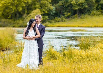 A couple stands together in a grassy meadow by a serene lake at a charming Leesburg VA wedding venue. The woman in a white floral gown smiles as the man in a dark suit gently embraces her from behind. Trees and greenery fill the background under a clear sky.