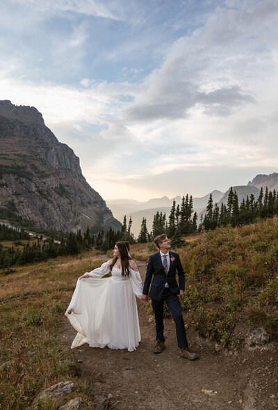 A bride and groom walk along a mountain trail surrounded by evergreens and soft evening light during their Glacier National Park elopement, captured by Sydney Breann Photography.