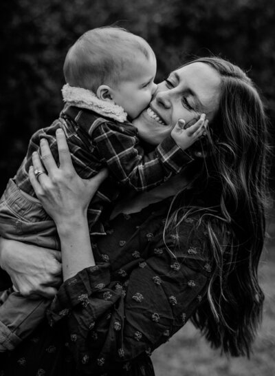 Black and white photo of baby boy twin kissing his mom on the cheek and holding her face while mom is smiling.