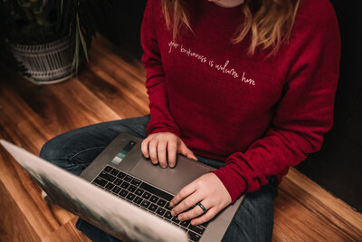 a women in a red sweater sits cross legged on the floor with her open laptop resting on her legs