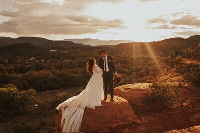 A couple stands on a red rock cliff, with one person holding a bouquet, as they gaze into each other's eyes. The sunset casts a golden light over the dramatic landscape, enhancing the romantic and majestic feel of the moment.