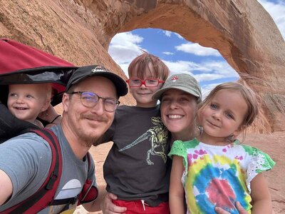 A fun and adventurous family moment captured by an Albuquerque family photographer. This image features a family selfie while canoeing on the river, with everyone wearing red life jackets. The man carries a baby as they sit together in a blue and gray canoe, showcasing a joyful and memorable experience in nature.