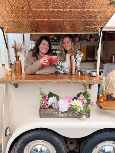 Owners of Sol & Spirits pose inside finished horse trailer bar with cocktails in hand in a "Cheers" pose. The bar window is open and the bar is decorated with flowers.