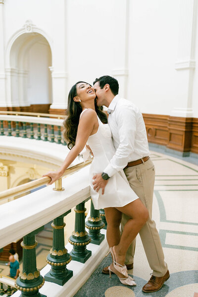 Man kissing woman beside a large railing in an ornate building