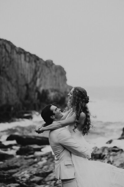 Black and white photo of bride and groom laughing with ocean cliff in background