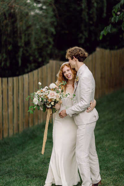 Peyton Curry and her husband share a joyful, candid moment while holding each other during their intimate backyard wedding portraits in Richmond, Virginia. With her floral bouquet in hand, this film-style wedding photography captures the beauty of documentary-style storytelling.