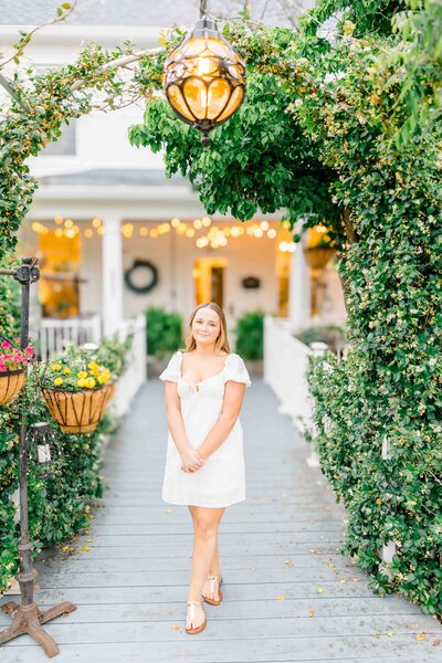 A woman in a white dress stands on a wooden pathway adorned with greenery and hanging plants. In the background, the charming white house with its veranda and string lights evokes the artistry of a skilled senior photographer from Fayetteville, NC. A decorative lantern hangs above her.