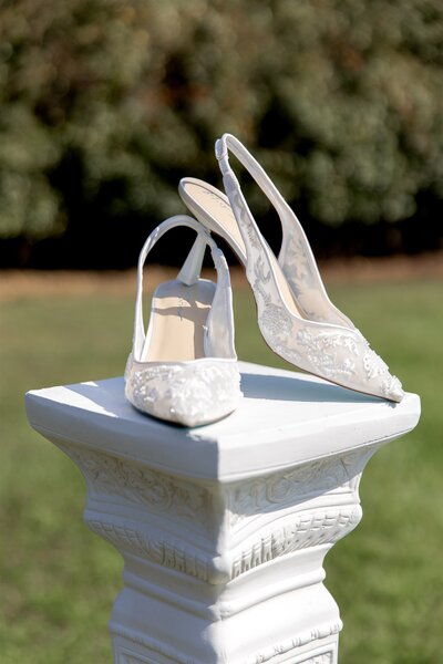 Close-up detail of white bridal heels on a pedestal at outdoor North Carolina wedding, captured in fine-art photo and video style.
