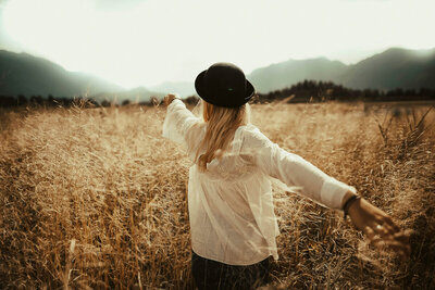 Woman with long blonde hair wearing a cream lace-accented shirt and black felt hat, walking through a golden wheat field with arms open wide, mountains in the background, symbolizing freedom, joy, and acceptance in Christ.”