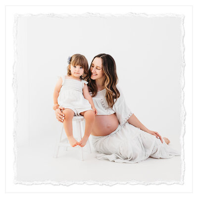 The photo shows a beautiful moment between a pregnant mom and her three-year-old daughter. The mom, dressed in a flowing white gown, sits on the floor, gently embracing her daughter, who wears a matching white ensemble. The setting is an all white backdrop, creating a clean and classic feel. The photograph was captured by talented photographer Bri Sullivan.