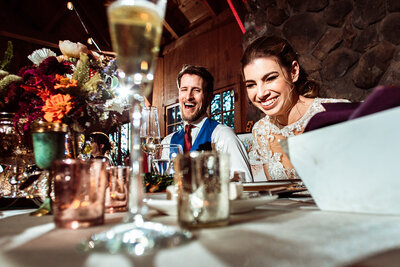 Newlyweds seated for champagne toast laugh during bridal party speech.