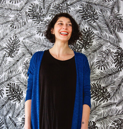 A portrait of artist and textile designer Skye McNeill. Skye wears a blue cardigan and a black shirt and stands in front of a wall sized block print of palm leaves. Photograph by Beth Olsen Creative.