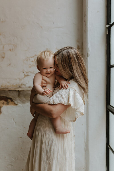 Black and white image of Kaz and her youngest child.  Kaz is sitting on the floor in a flowing white dress, lifting her baby up on her arms above her.  They are both smiling at each other. 