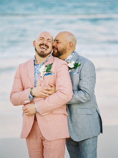 Two grooms embracing and laughing together on the beach during their destination wedding, at Paradisus Palma Real Resort captured in natural light by Asia Pimentel Photography.