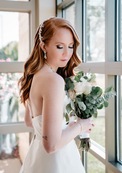 a bride standing by a window holding flowers and looking down at her shoulder