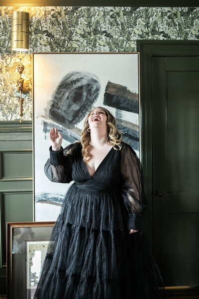 Ashleigh C. Henry, life coach, writer, speaker, and author, laughing joyfully at the Blind Tiger Asheville near a piece of art, wearing a black ballgown, in an olive green room with crown molding and wallpaper. 
