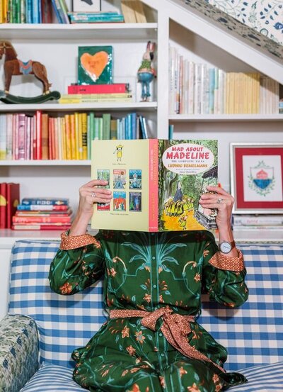 Woman in green patterned dress sitting on a blue check couch in front of a bookcase holding up a book in front of her face