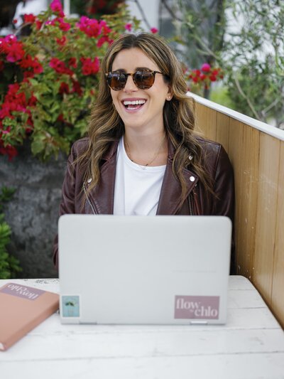 A woman sitting wearing sunglasses at an outdoor table with her laptop and notebook, smiling and looking away from the camera