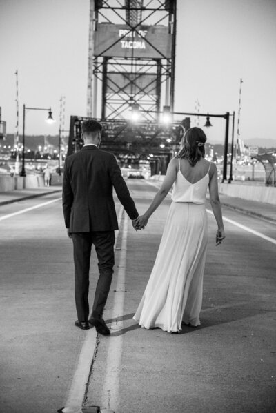 Couple wearing a suit and simple wedding dress, walking away from the camera down the middle of a closed bridge in downtown Tacoma