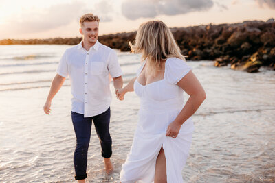 Woman in white dress holding man's hand and running on the shore of a Tampa area beach during an engagement portrait session