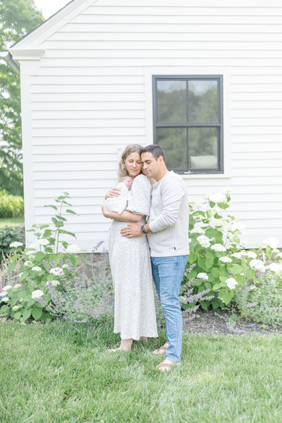 A husband and wife sit closely together in a blooming spring garden, lovingly holding their newborn. The mother cradles the baby in her arms while the father gently leans in, both gazing at their child with warmth and joy. Soft sunlight filters through the flowers and trees, adding to the serene and tender atmosphere.
