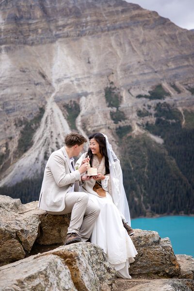 A couple in wedding attire sitting on the cliffs of Peyto Lake sharing a wedding cake 