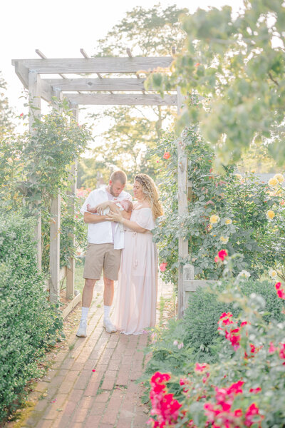 Mom sitting on a patio, gently holding her swaddled newborn in her arms, gazing down at him with a loving expression. Dad stands behind her, looking over her shoulder with a proud and tender gaze, creating a warm family moment outdoors