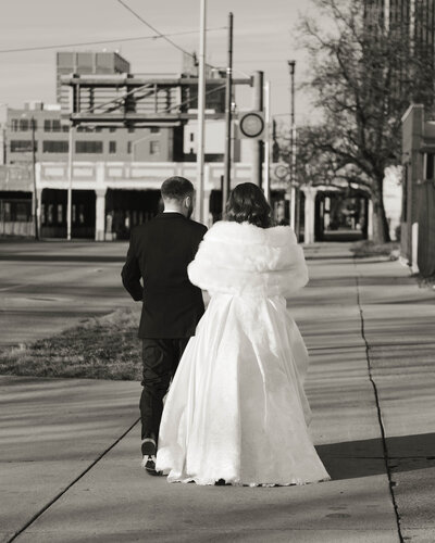 a black and white image of a bride and groom with linked arms walking down the street from behind