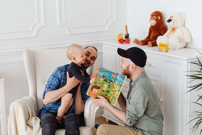 Two dads cozying up to read a story to their baby, showcasing a warm, nurturing family moment centered around the joy of shared storytelling.