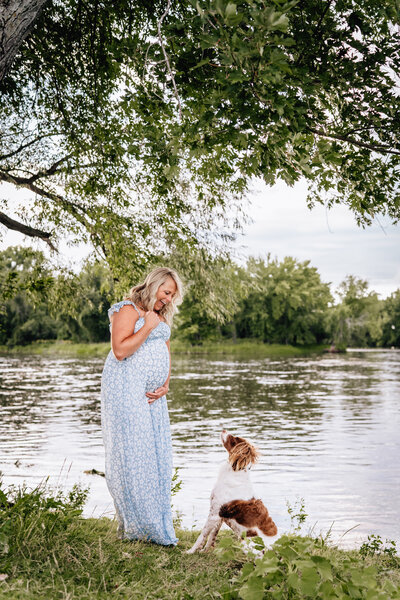 A pregnant woman in a light blue floral dress stands by a serene lake, looking down with a joyful smile at her dog, who is sitting attentively at her feet. The background is filled with green trees and a calm water surface.