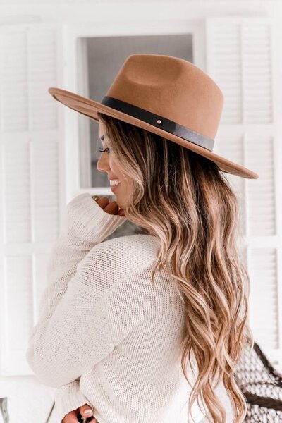 A woman with long, wavy hair is wearing a beige wide-brimmed hat and a white sweater. She is smiling and looking to the side, embodying the elegance of branding and Showit web design for salons. White shutters are in the background.