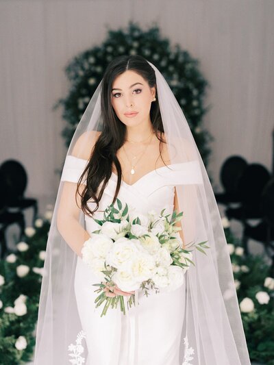 Elegant bride holding a lush white bridal bouquet, standing gracefully amidst beautiful green and white floral aisle decorations, showcasing exquisite ceremony design by a Virginia wedding florist.