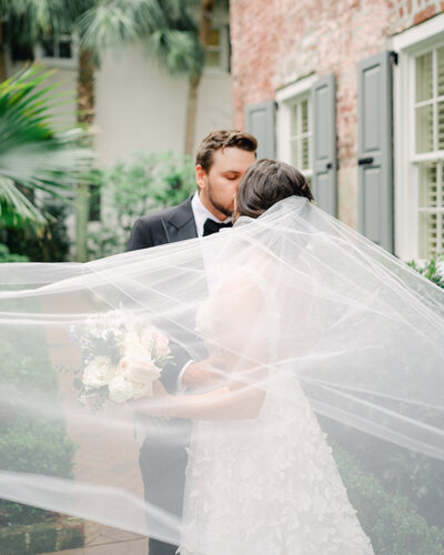 Bride and Groom kiss while the bridal veil blows in the wind.  Portrait by Charleston Wedding Photographer, Sarah Woods.