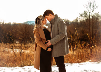A pregnant woman and a man are standing in a snowy area with tall brown grass, trees, and mountains behind them. They're touching heads with their eyes closed with all hands on moms growing belly.