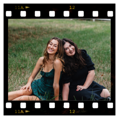 Clarae and Taressa, wedding and event planners in Oregon, sit in the grass smiling with their backs against each other. 