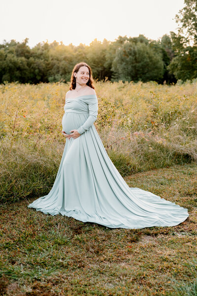 Pregnant mom wearing green holding her belly and looking over her shoulder smiling in nature park in Eastern Shore Maryland.