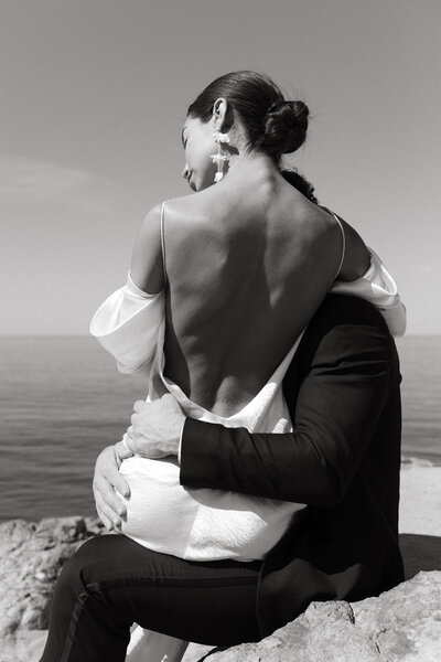 black and white photo of bride on grooms lap cliffside