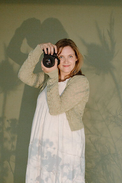 Coeur d'Alene photographer Brittany Barb in a white dress and tan cardigan holding camera against green wall with artistic shadow