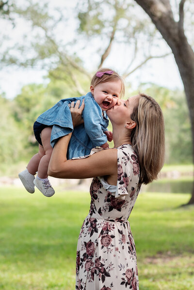 Mom in floral dress holds giggling baby girl in air and kisses her in Atlantic Beach, FL.