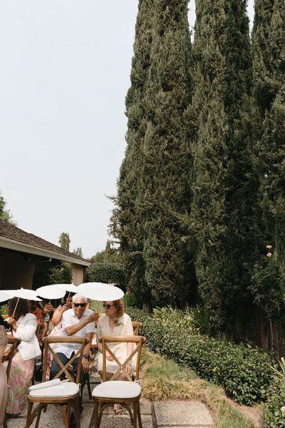 Carmel, CA backyard wedding. Wedding guests sitting at ceremony with parasols in the summer sun