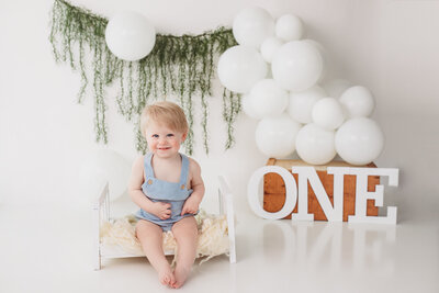 Toddler boy sitting on a miniature bed smiling. There are while balloon and greenery surrounding him.