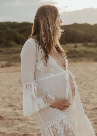 Pregnant woman in white lace robe posing for a maternity photoshoot at the beach so she has memories of this life-changing time.