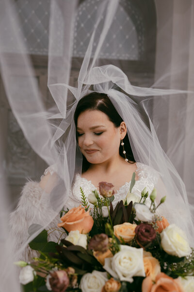 Wedding Photography of dark haired bride in a vintage lace gown and grey fur jacket draped below her shoulders, looking down smiling with her veil blowing up above her and around her, holding a gorgeous and large bouquet of moody, vintage flowers, standing in front of an old church door in Milwaukee, WI